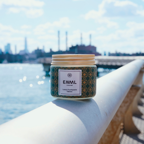 A jar of ENML Luxury Toothpaste Tablets placed on a railing with a scenic waterfront and city skyline in the background, highlighting its portability and eco-friendly design.